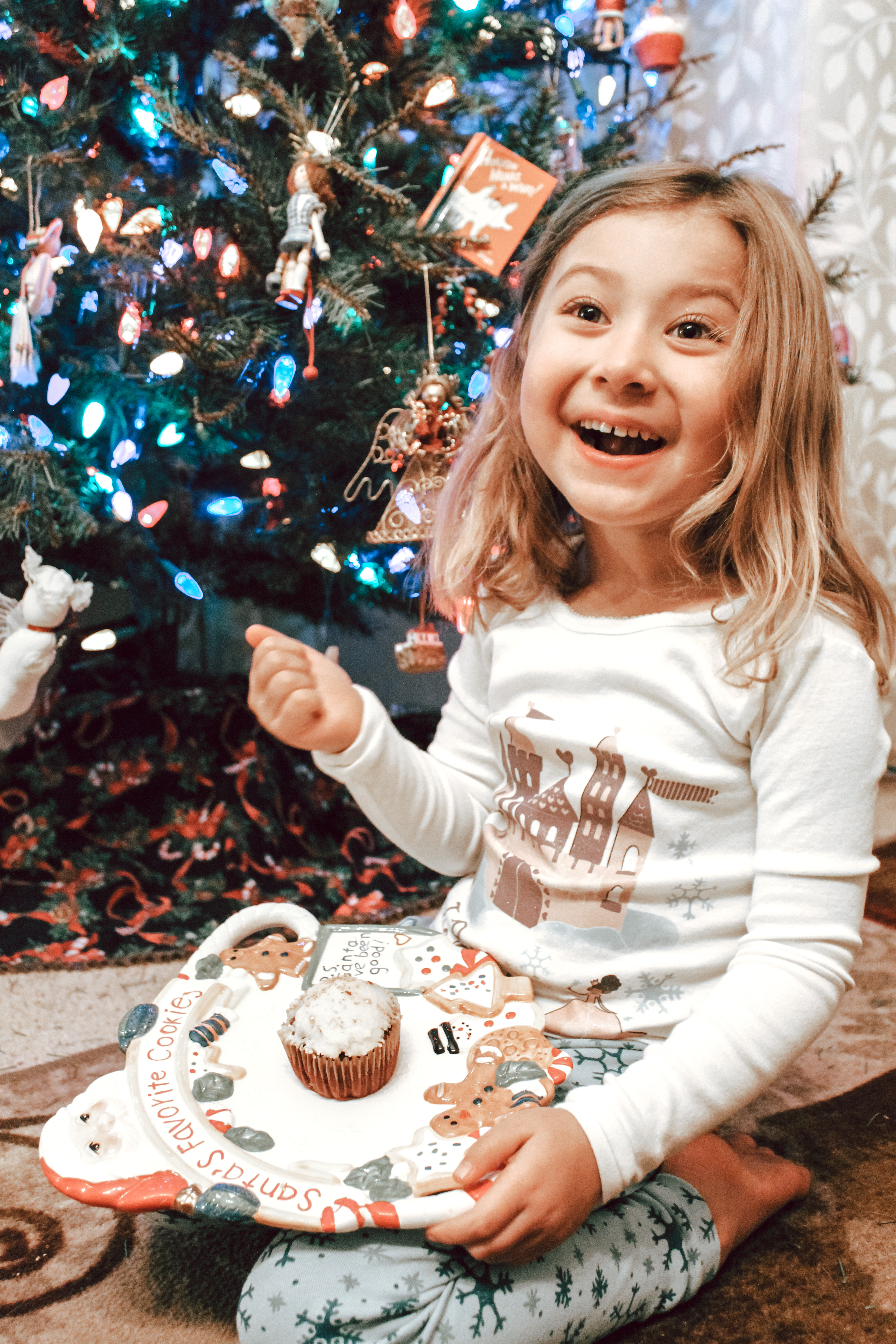 A little girls sits in front of a decorated Christmas tree with a cupcake for Santa Claus