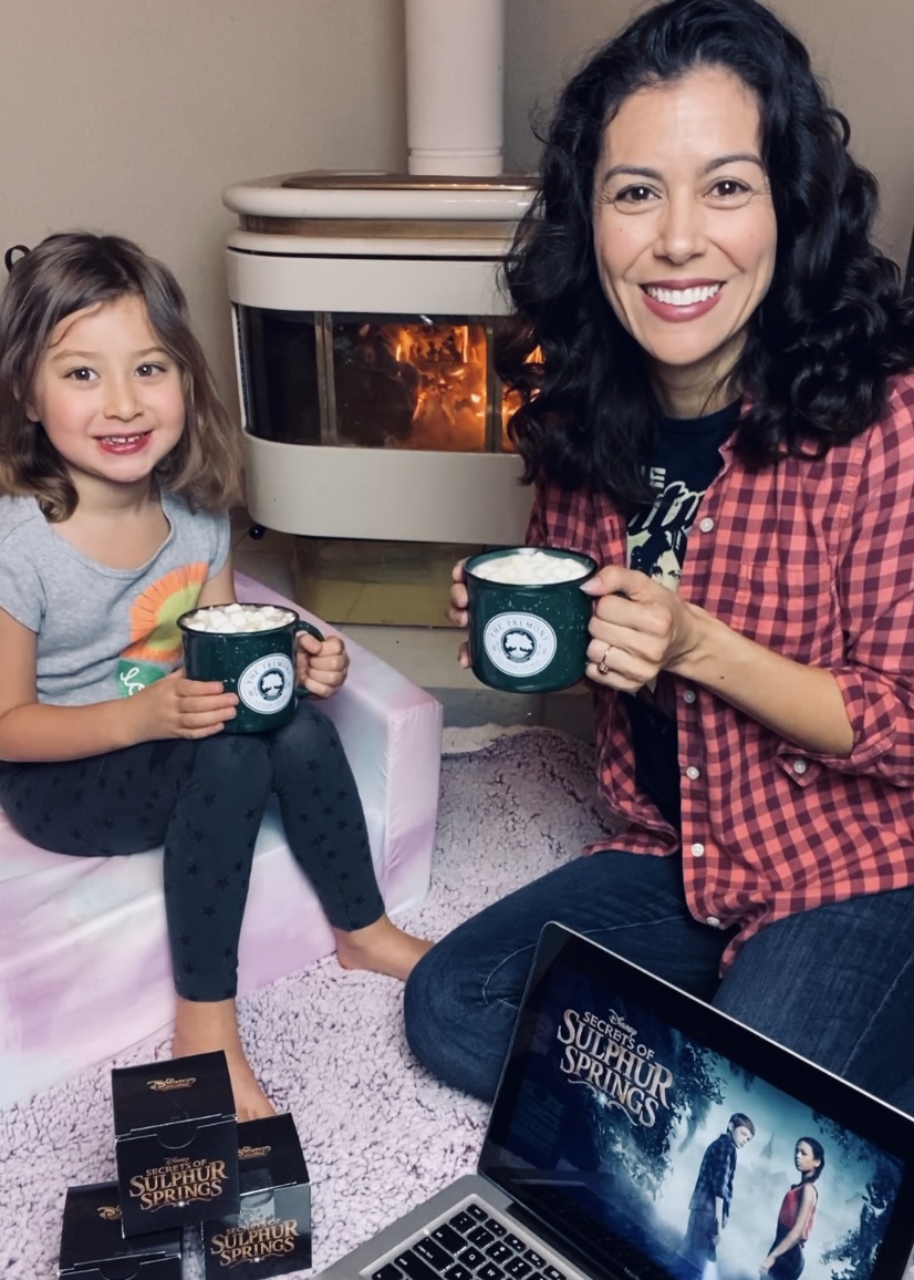 Casey Dacanay and daughter Olivia sit by the fire sipping hot cocoa out of green camp mugs featuring The Tremont, a fictional hotel from the Disney Channel television show Secrets Of Sulphur Springs